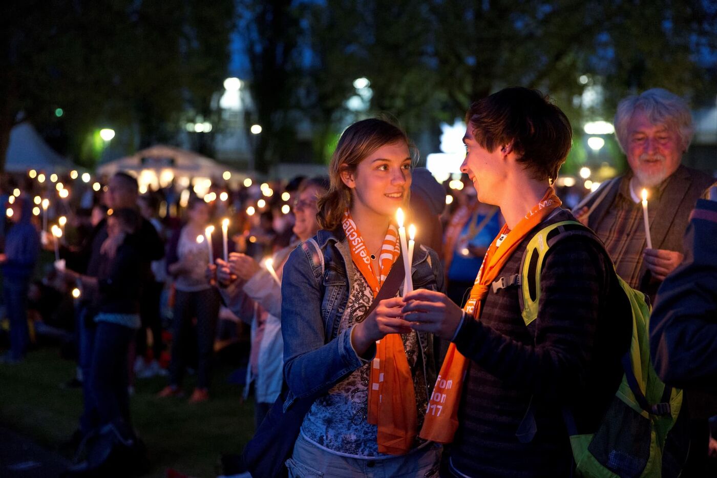 eine Gruppe von Menschen steht abends mit leuchtenden Kerzen in der Hand in einer Stadt. Sie lächeln.